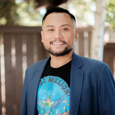 Huy Tran, a man with a black beard and short hair, stands in front of a fence and trees. He is wearing a dark blue blazer over a shirt with a blue circle that says 11 million! with a hummingbird in mid-flight in the middle.