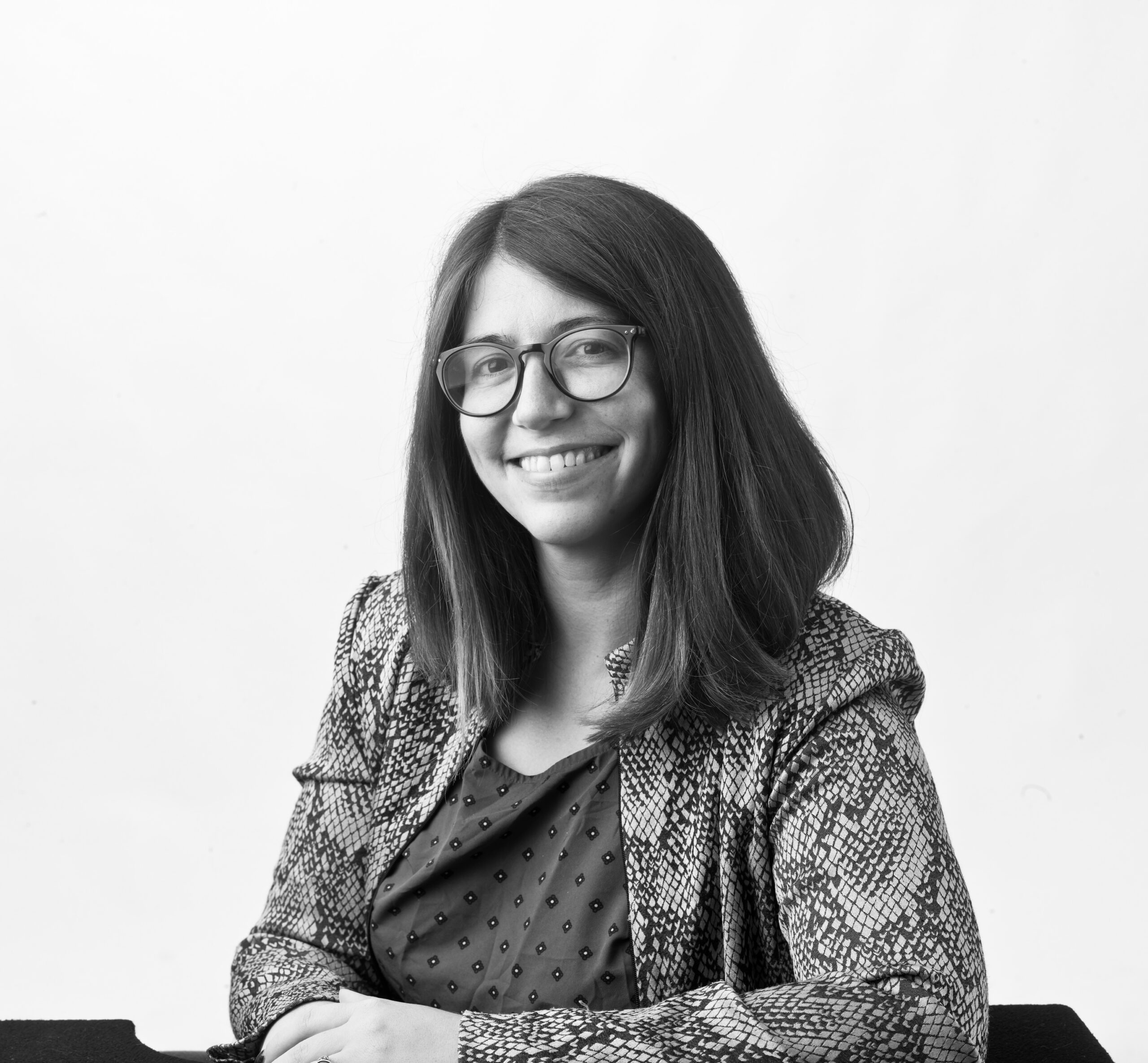 Abbee Cox, a person with shoulder-length straight dark hair, poses in front of a white background. She is wearing a patterned blazer and spotted shirt. The photo is in black and white.