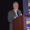 Anthony Prince, a white man with a grey and white beard and hair, wearing glasses and a grey suit, stands at a podium. The podiium has a purple and yellow SEIU banner on it.