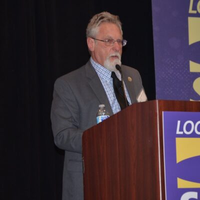 Anthony Prince, a white man with a grey and white beard and hair, wearing glasses and a grey suit, stands at a podium. The podiium has a purple and yellow SEIU banner on it.