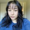 Itzel Romero, a person with medium-length wavy black hair and bangs, looks at the camera. They are wearing a bright blue sweater.