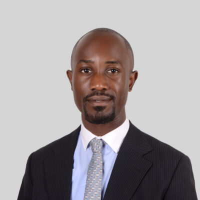 Henry Byansi, a bald Black man with a goatee and mustache, stands in front of a grey background. He is wearing a black blazer, white collared shirt, and grey patterned tie.