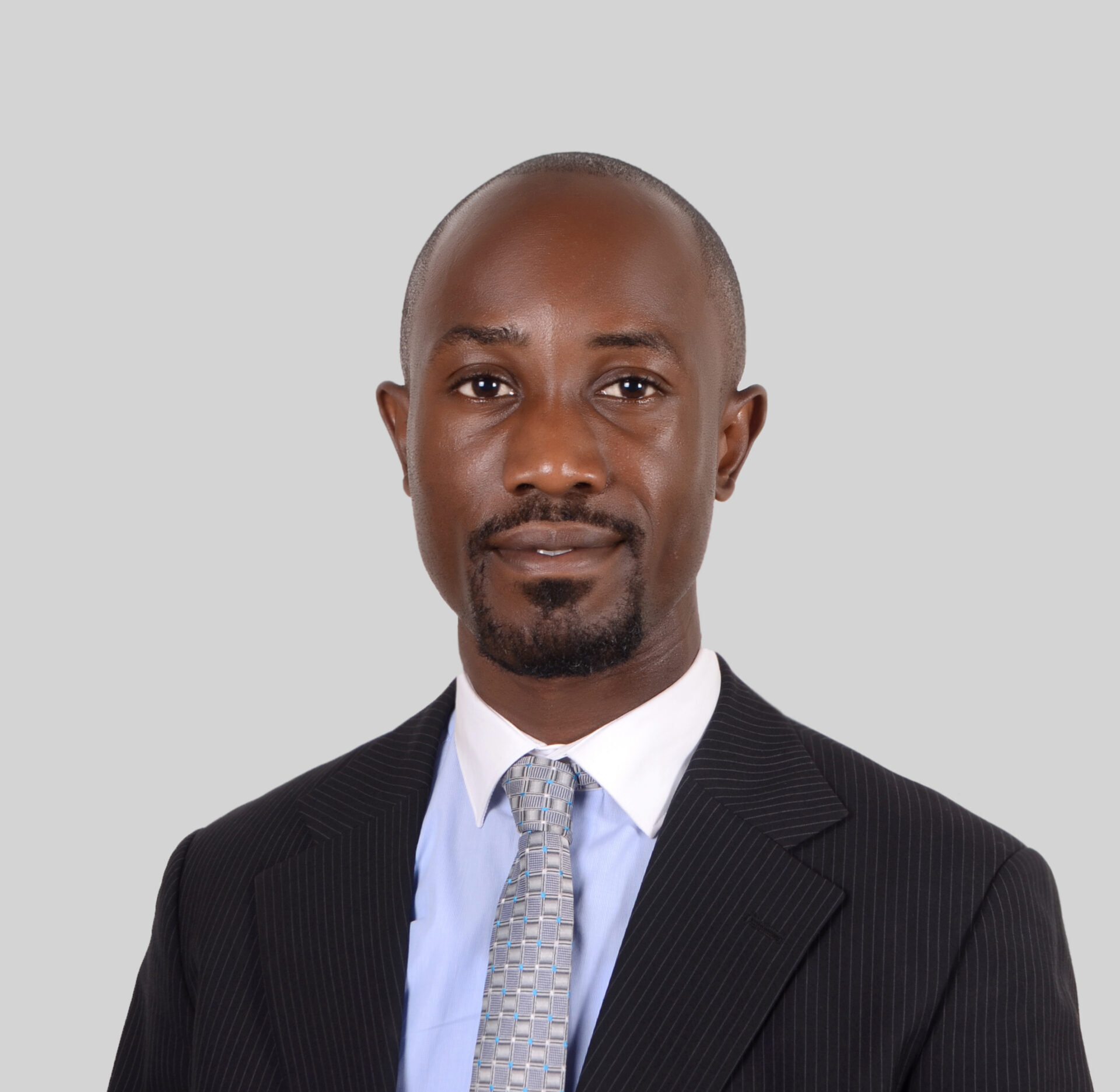 Henry Byansi, a bald Black man with a goatee and mustache, stands in front of a grey background. He is wearing a black blazer, white collared shirt, and grey patterned tie.