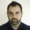 Omar Shakir, a person with cropped dark brown hair and a light beard and moustache, is wearing a dark collared shirt. He looks at the camera without smiling.