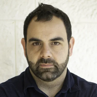 Omar Shakir, a person with cropped dark brown hair and a light beard and moustache, is wearing a dark collared shirt. He looks at the camera without smiling.