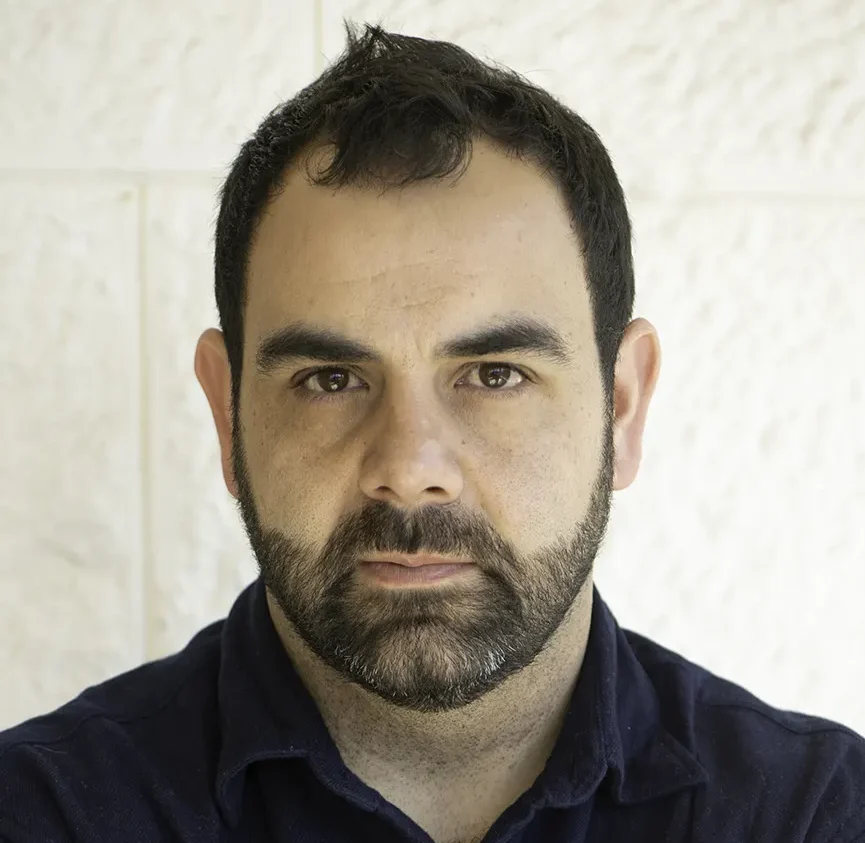Omar Shakir, a person with cropped dark brown hair and a light beard and moustache, is wearing a dark collared shirt. He looks at the camera without smiling.