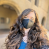 Sierra Maciorowski, a white person with long wavy dark brown hair, looks at the camera. She is wearing a black KN95 mask, a bright blue shirt, and a black netted sweater. In the background, you can see the arches of Stanford Law School and windows.