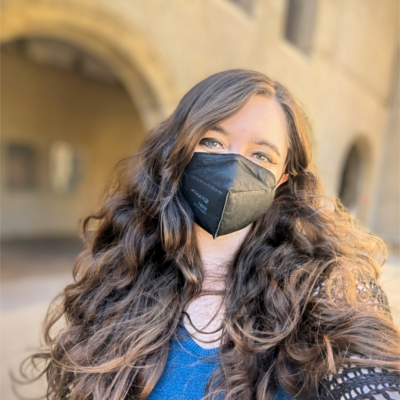 Sierra Maciorowski, a white person with long wavy dark brown hair, looks at the camera. She is wearing a black KN95 mask, a bright blue shirt, and a black netted sweater. In the background, you can see the arches of Stanford Law School and windows.