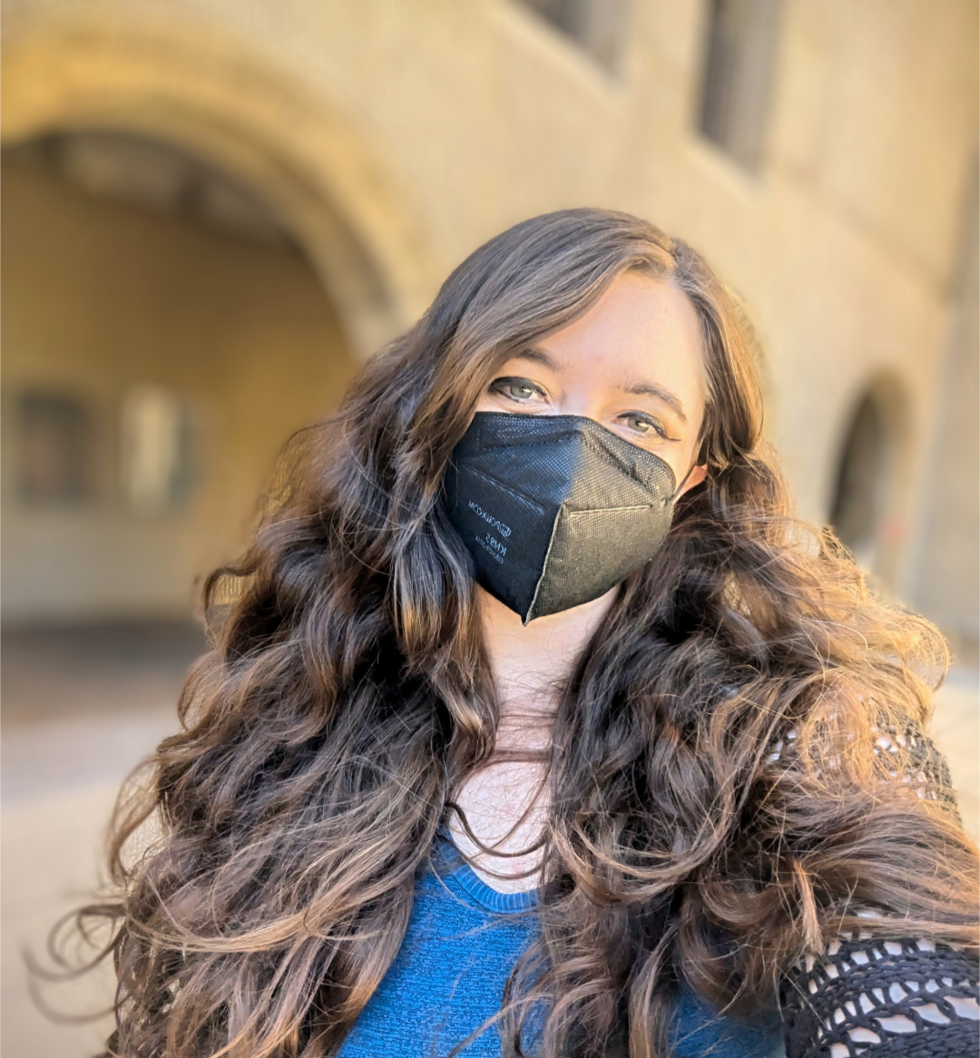 Sierra Maciorowski, a white person with long wavy dark brown hair, looks at the camera. She is wearing a black KN95 mask, a bright blue shirt, and a black netted sweater. In the background, you can see the arches of Stanford Law School and windows.