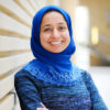 Shirin Sinnar poses smiling in front of a beige wall. She is wearing a blue shirt and brighter blue hijab.