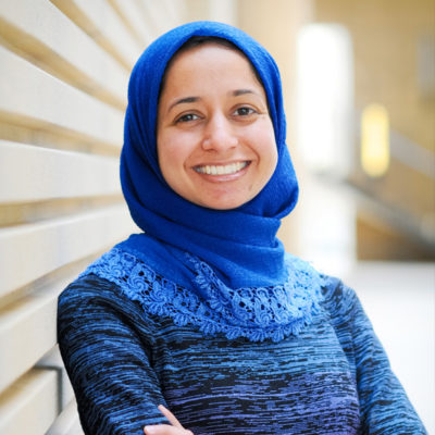 Shirin Sinnar poses smiling in front of a beige wall. She is wearing a blue shirt and brighter blue hijab.