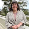 Tori Porell, a white person with her hands clasped in front of her, smiles at the camera. She has shoulder length brown hair and bangs, wears glasses, and has a grey patterned blazer.