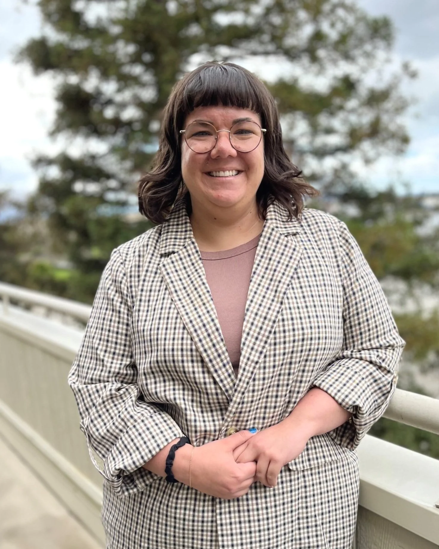 Tori Porell, a white person with her hands clasped in front of her, smiles at the camera. She has shoulder length brown hair and bangs, wears glasses, and has a grey patterned blazer.