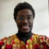 Bukongo, a Black man with short hair and round glasses, smiles at the camera. He has a faint moustache and beard, and is wearing a red and yellow patterned shirt.
