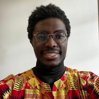 Bukongo, a Black man with short hair and round glasses, smiles at the camera. He has a faint moustache and beard, and is wearing a red and yellow patterned shirt.