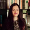 Pantea Javidan, a person with long brown hair wearing a patterned shirt and jacket, poses in front of bookshelves.