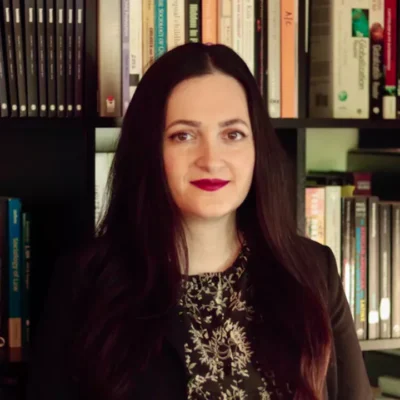 Pantea Javidan, a person with long brown hair wearing a patterned shirt and jacket, poses in front of bookshelves.