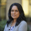 Jayashri Srikantiah poses in front of blurred beige walls and green shrubs. She is smiling softly, with slightly wavy black hair, wearing a grey sweater and blue pattened shirt.