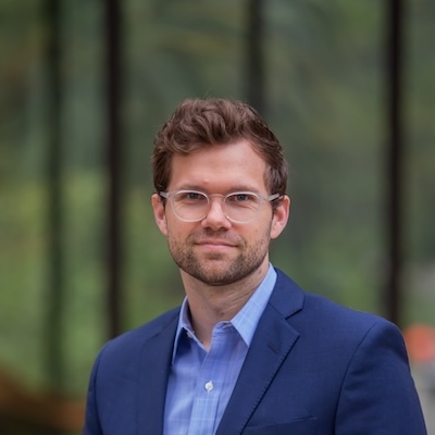 Shaw Drake, a man with medium-brown hair, a short beard, and glasses, looks at the camera with a faint smile. He is wearing a navy blue suit jacket and blue collared shirt.
