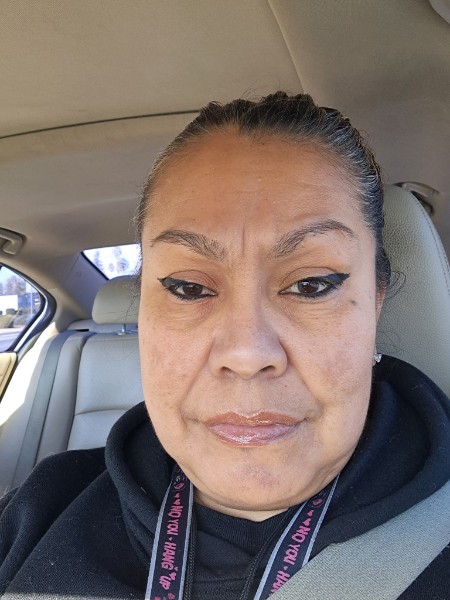 Nancy Torres, a woman with brown hair pulled back, looks at the camera in a selfie. She is sitting in a car with grey seats, and wearing a dark colored jacket.